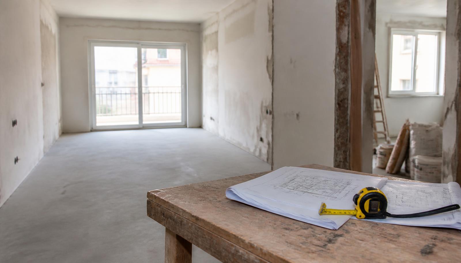 Interior de apartamento português em obra, com paredes rebocadas, betonilha nova e plantas de projeto com fita métrica sobre uma bancada de madeira